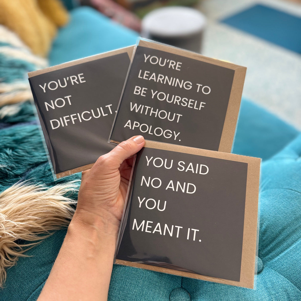 Three black square cards with white text are held in a hand over a teal tufted sofa. The cards read: 'YOU'RE NOT DIFFICULT.' 'YOU SAID NO AND YOU MEANT IT.' 'YOU'RE LEARNING TO BE YOURSELF WITHOUT APOLOGY.'