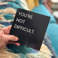 A hand holds a black square card with the text 'YOU'RE NOT DIFFICULT.' in white sans-serif lettering, set against a cozy background of a blue and teal faux fur blanket.