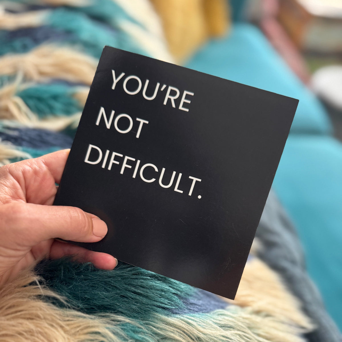 A hand holds a black square card with the text 'YOU'RE NOT DIFFICULT.' in white sans-serif lettering, set against a cozy background of a blue and teal faux fur blanket.