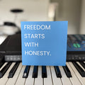 A square blue greeting card with the white text 'FREEDOM STARTS WITH HONESTY.' propped up on a piano keyboard, with a blurred indoor background and musical equipment behind it.