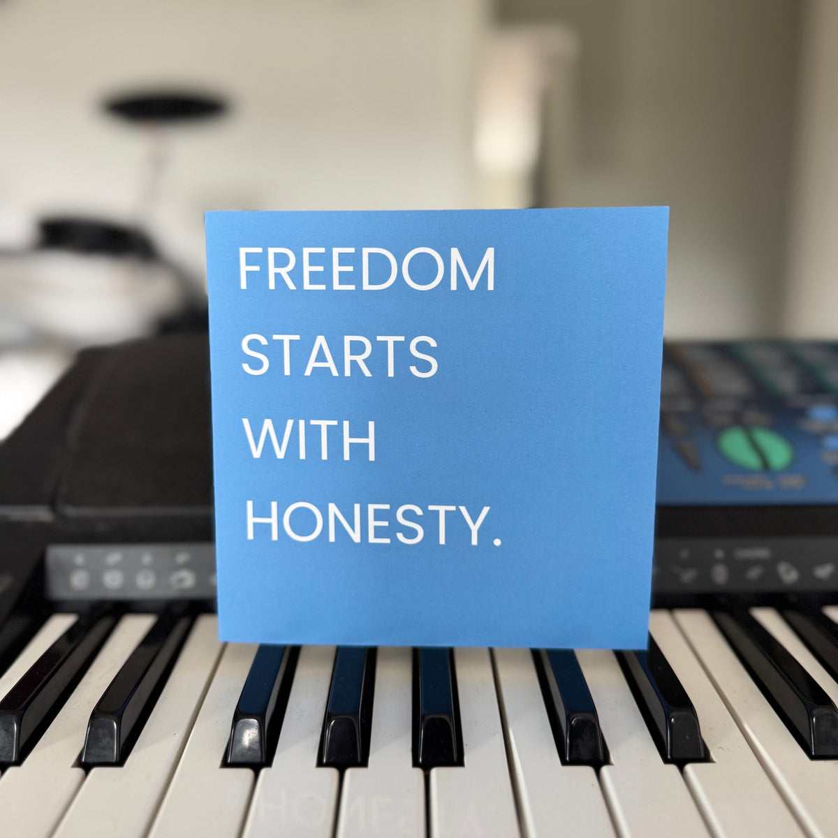 A square blue greeting card with the white text 'FREEDOM STARTS WITH HONESTY.' propped up on a piano keyboard, with a blurred indoor background and musical equipment behind it.