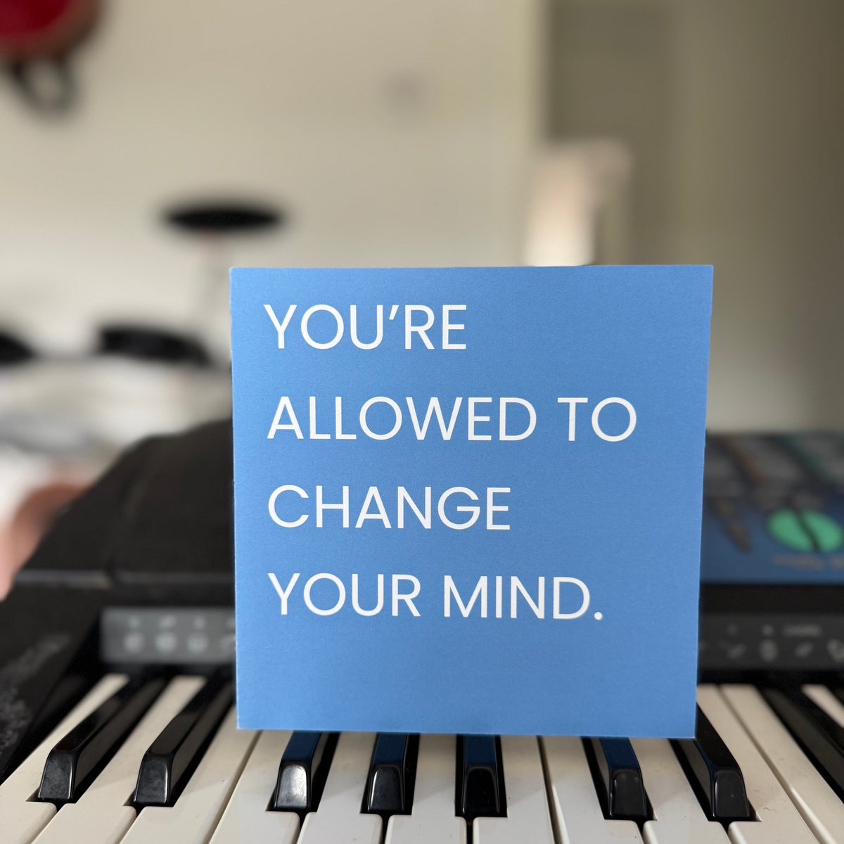 A blue square greeting card with white text reading 'YOU'RE ALLOWED TO CHANGE YOUR MIND.' placed on a keyboard in a real indoor setting with a blurred background.