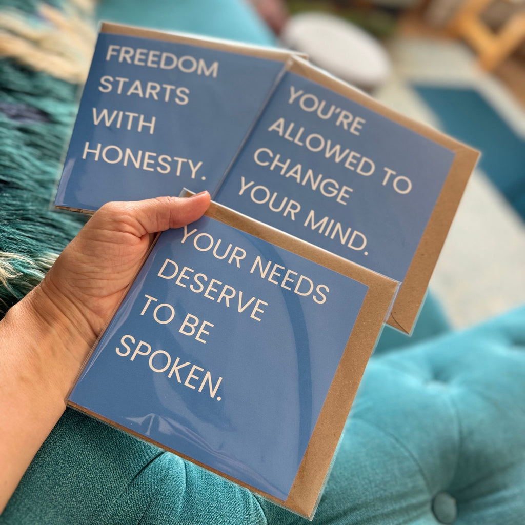A hand holds three blue greeting cards with quotes against a teal tufted sofa background. The cards have a matte blue finish with white text and a brown kraft paper border.