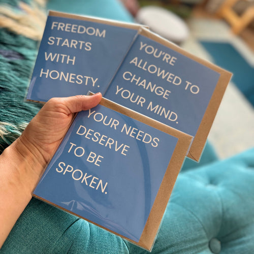 A hand holds three blue greeting cards with quotes against a teal tufted sofa background. The cards have a matte blue finish with white text and a brown kraft paper border.