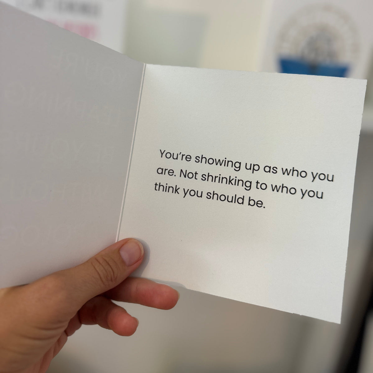 A hand holding an open greeting card with a printed message on the inside: 'You're showing up as who you are. Not shrinking to who you think you should be.