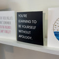 A black card with white text reading 'YOU'RE LEARNING TO BE YOURSELF WITHOUT APOLOGY.' displayed on a white shelf next to other decorative signs.