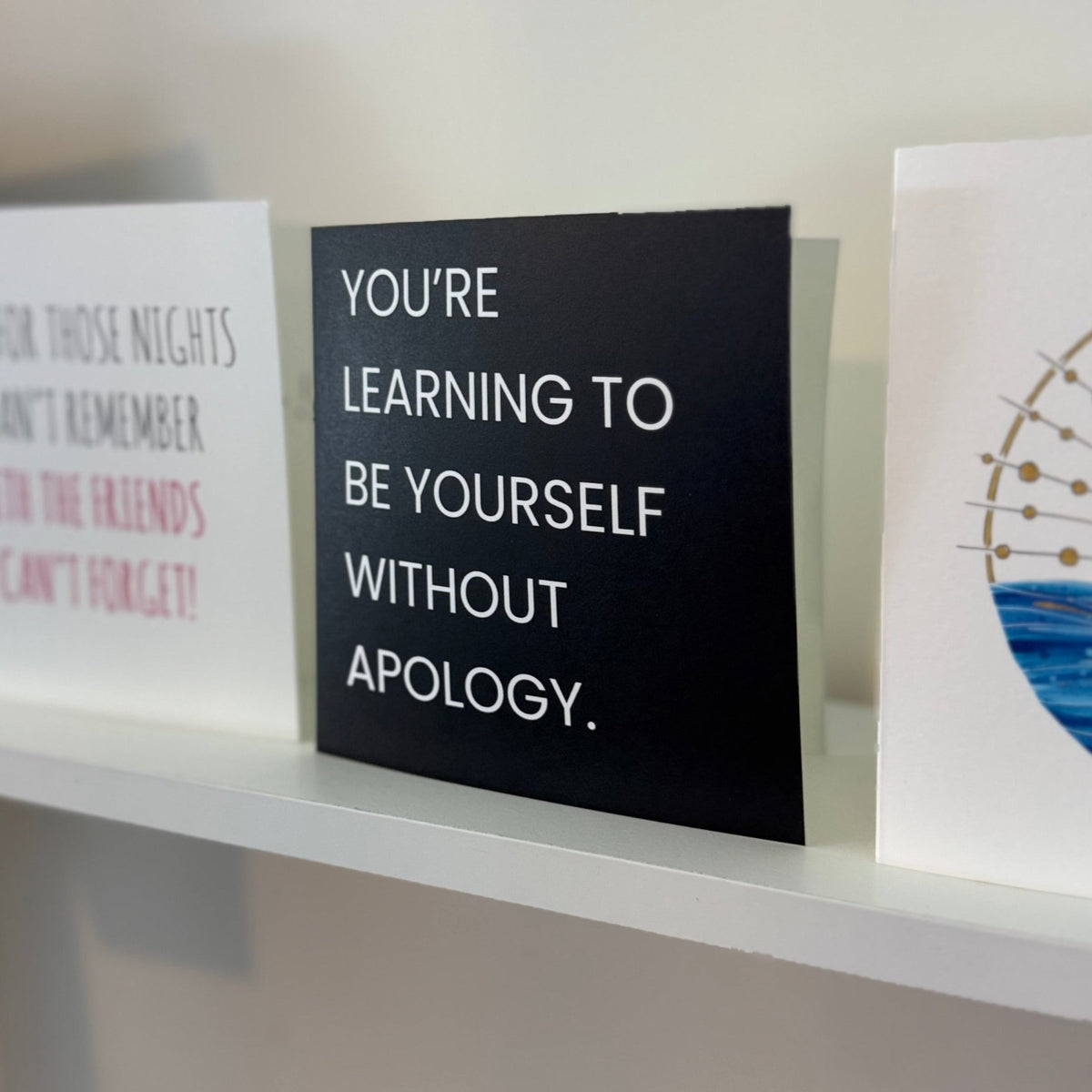 A black card with white text reading 'YOU'RE LEARNING TO BE YOURSELF WITHOUT APOLOGY.' displayed on a white shelf next to other decorative signs.