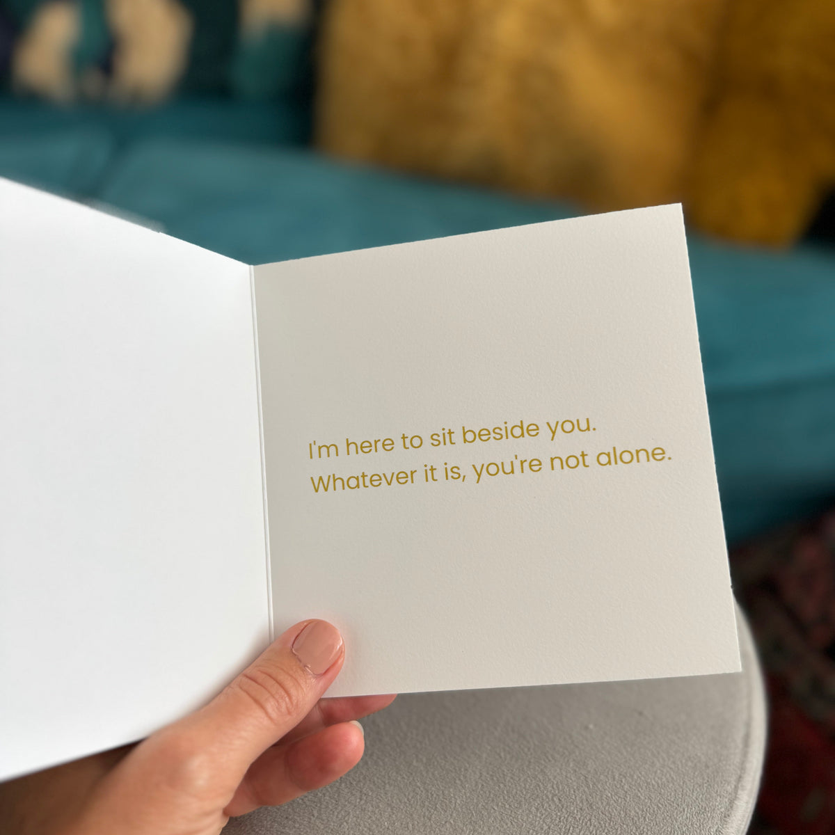 A hand holds an open white greeting card with gold foil text that reads, 'I'm here to sit beside you. Whatever it is, you're not alone.