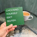 A hand holds a green square card with the text 'CHOOSING YOURSELF ISN'T SELFISH.' in white capital letters, with a blurred background showing a cup of coffee and a green wooden wall.