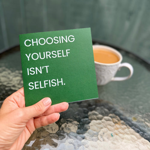 A hand holds a green square card with the text 'CHOOSING YOURSELF ISN'T SELFISH.' in white capital letters, with a blurred background showing a cup of coffee and a green wooden wall.