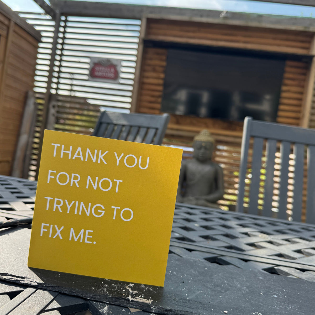 A square yellow card with white text reading 'THANK YOU FOR NOT TRYING TO FIX ME.' placed on a black metal patio table in an outdoor patio setting with wooden furniture and a slatted wall background.