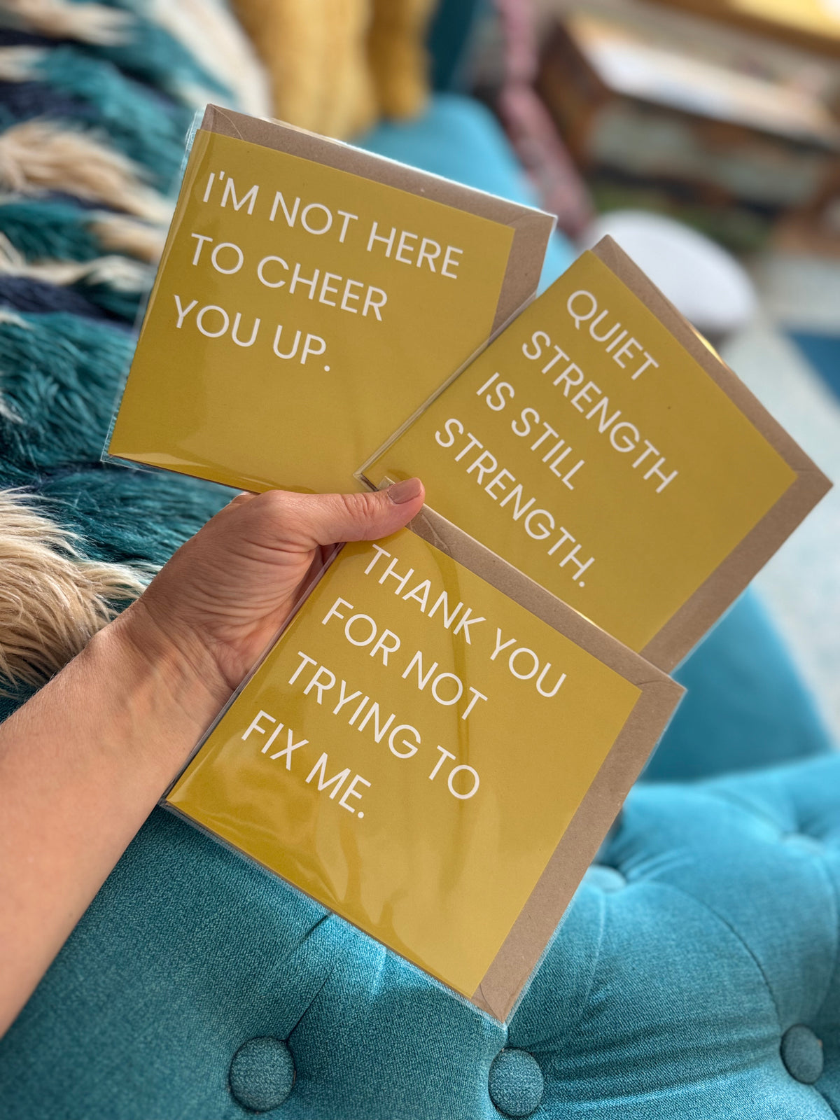 Three yellow greeting cards with white text are held in a hand against a teal tufted sofa. The cards read: 'I'M NOT HERE TO CHEER YOU UP.' 'QUIET STRENGTH IS STILL STRENGTH.' 'THANK YOU FOR NOT TRYING TO FIX ME.'