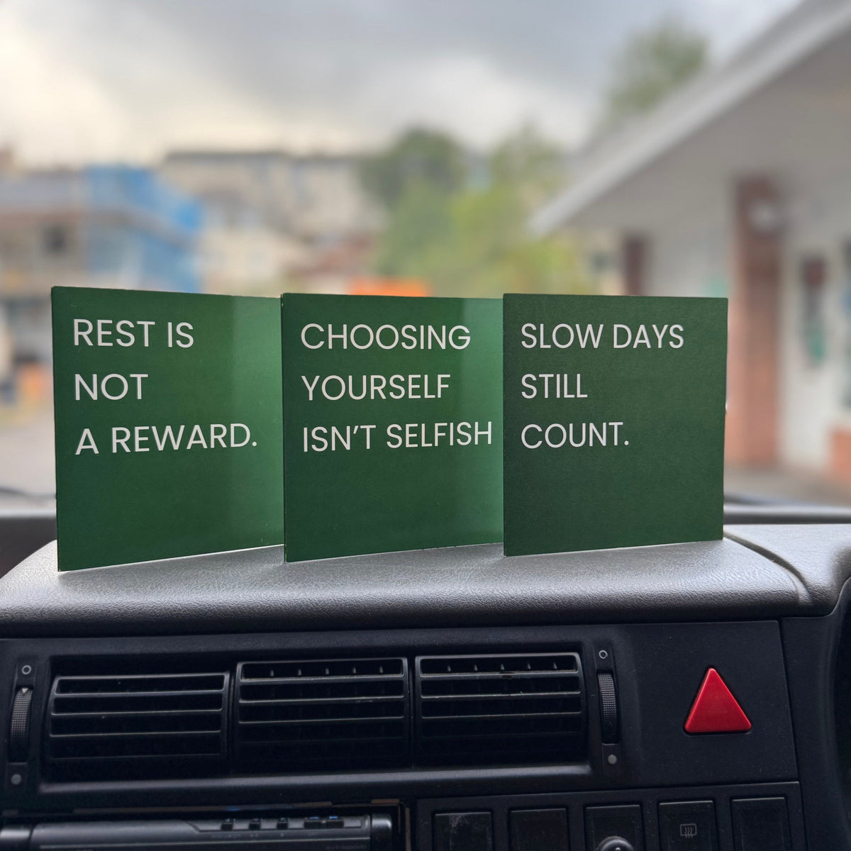 Three green square cards with white text displayed on a car dashboard. The signs read: 'REST IS NOT A REWARD.