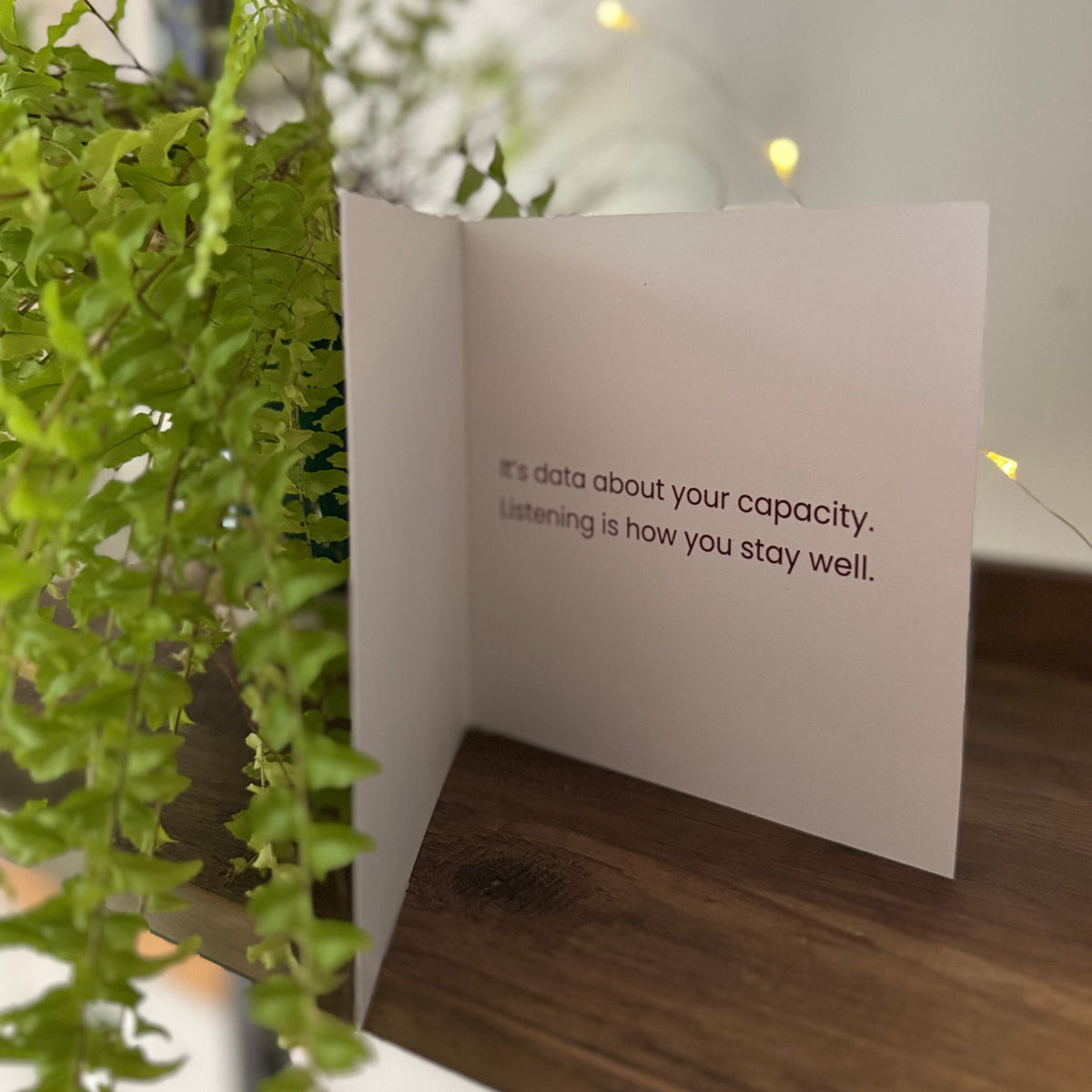 A white greeting card standing open on a wooden shelf, partially framed by a green trailing plant. The card has black text that reads, 'it's data about your capacity.