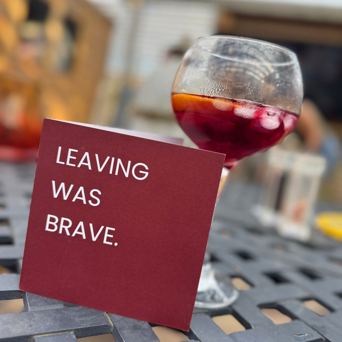 A red table card with the white text 'LEAVING WAS BRAVE.' standing on a black lattice table next to a glass of red cocktail in an outdoor patio setting.
