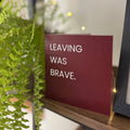A square red card with white text reading 'LEAVING WAS BRAVE.' displayed on a wooden shelf next to a green hanging plant and a black picture frame.