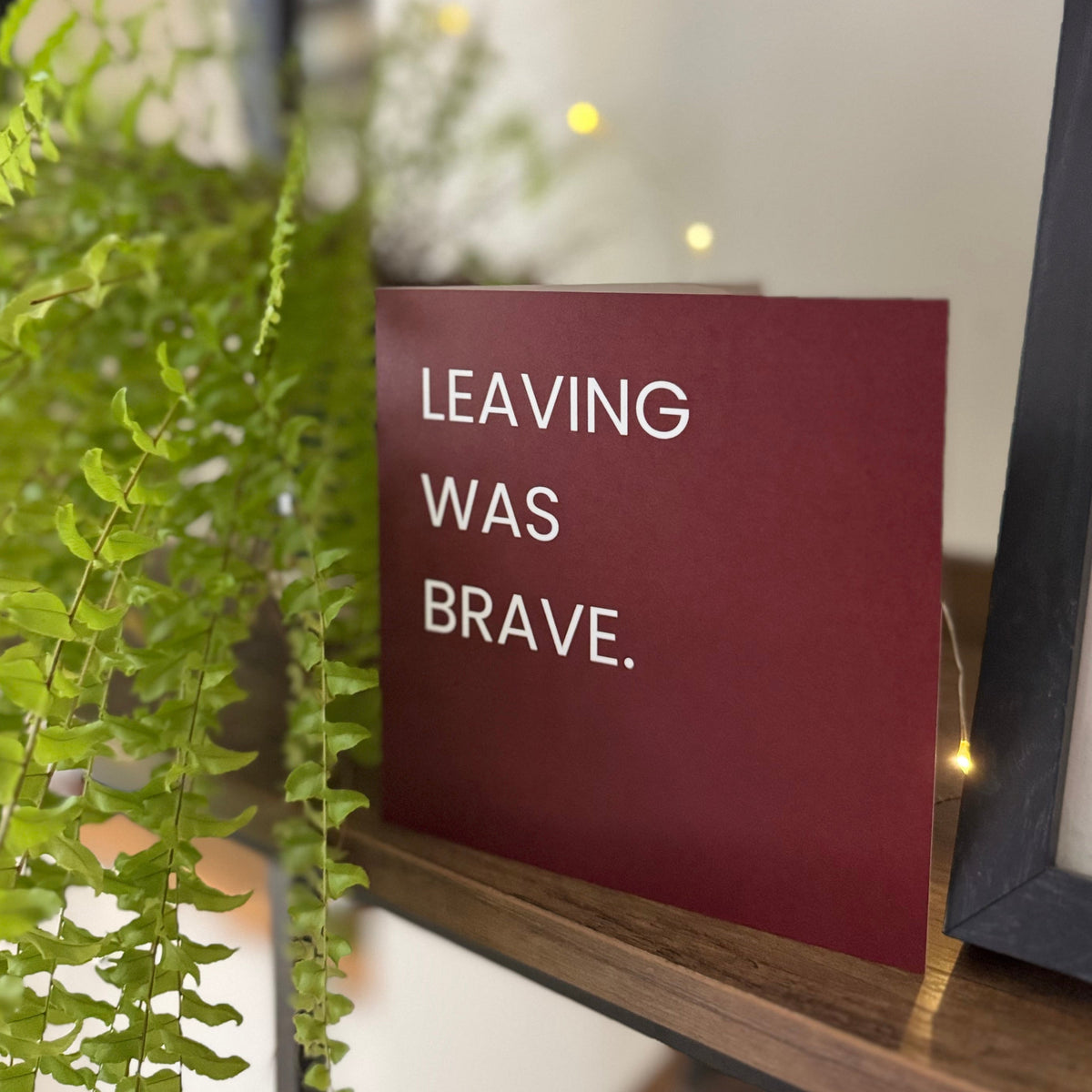 A square red card with white text reading 'LEAVING WAS BRAVE.' displayed on a wooden shelf next to a green hanging plant and a black picture frame.
