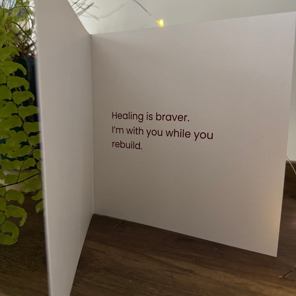 A white greeting card standing open on a wooden surface, printed with the message 'Healing is braver. I'm with you while you rebuild.