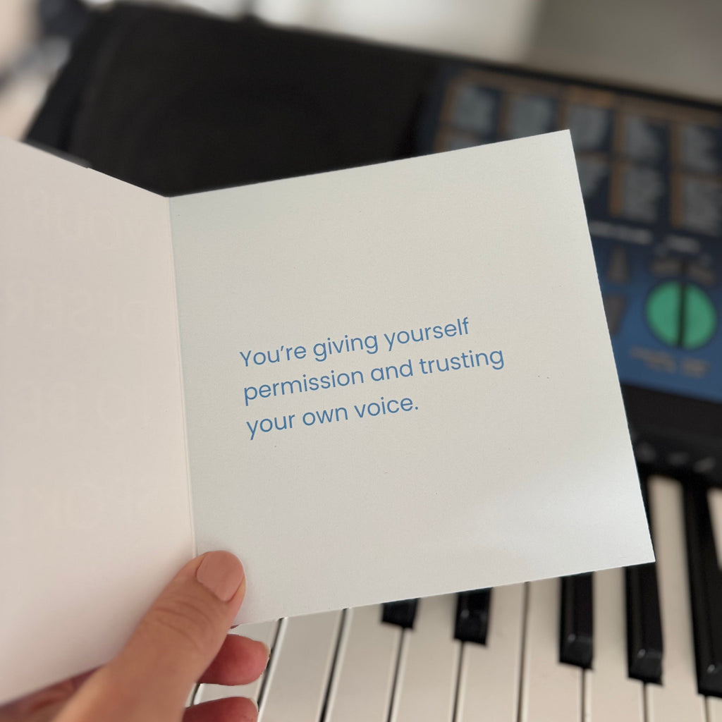 A hand holds an open white greeting card with blue text that reads, 'You're giving yourself permission and trusting your own voice.' in the foreground, with a blurred piano keyboard and room background behind it.