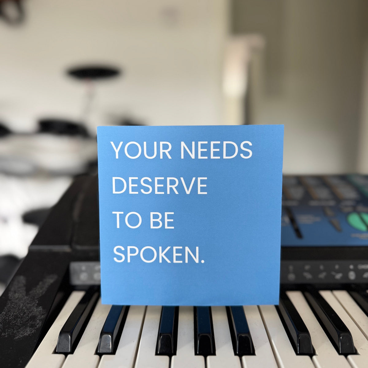 A blue square greetings card with white text reading 'YOUR NEEDS DESERVE TO BE SPOKEN.' placed on the keys of a keyboard in a blurred indoor setting.