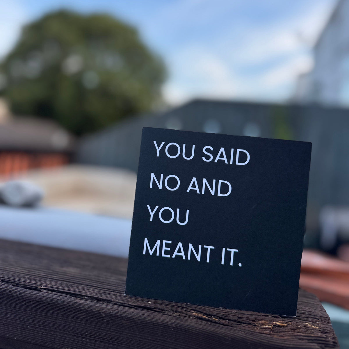A small black square sign with white text reading 'YOU SAID NO AND YOU MEANT IT.' standing on a dark wooden surface outdoors with a blurred blue sky, trees, and buildings in the background.