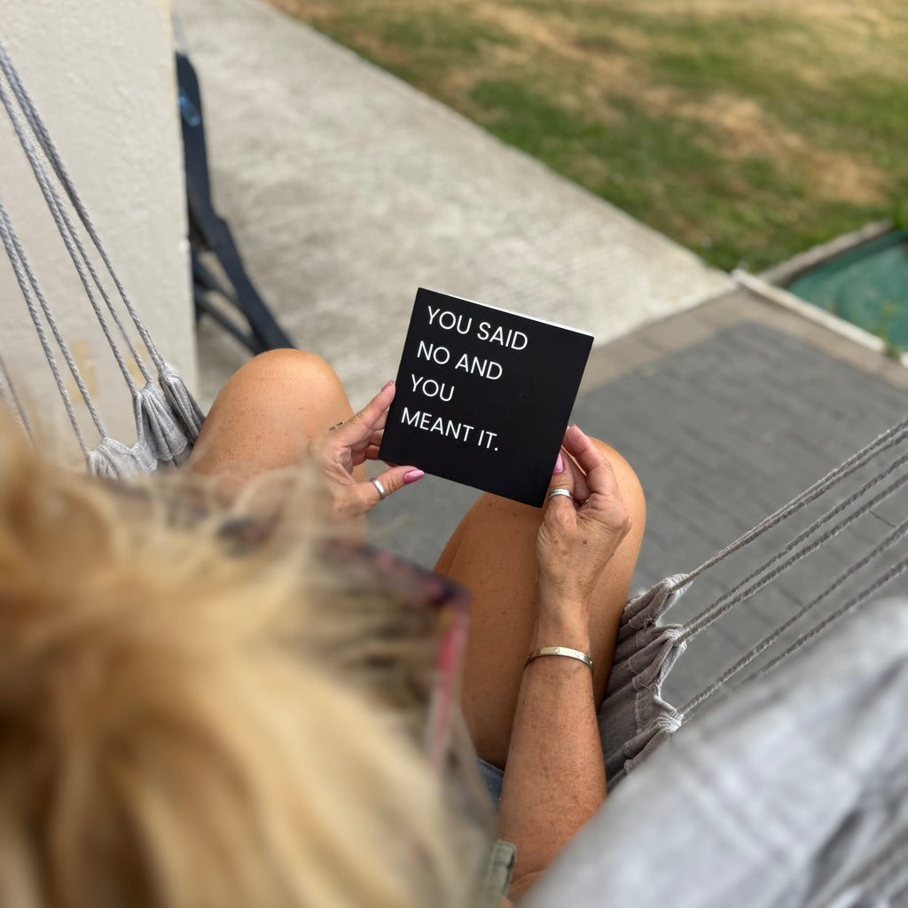 A person sitting in a hammock holds up a small black square sign with white text that reads, 'YOU SAID NO AND YOU MEANT IT.' The photo is taken from above, with the person's blonde hair in the foreground and a patio, grass, and pool visible in the background.