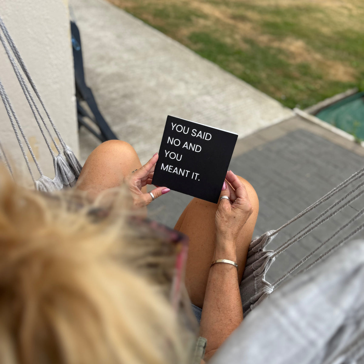 A person sitting in a hammock holds up a small black square sign with white text that reads, 'YOU SAID NO AND YOU MEANT IT.' The photo is taken from above, with the person's blonde hair in the foreground and a patio, grass, and pool visible in the background.