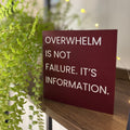A square red card with the text 'OVERWHELM IS NOT FAILURE. IT'S INFORMATION.