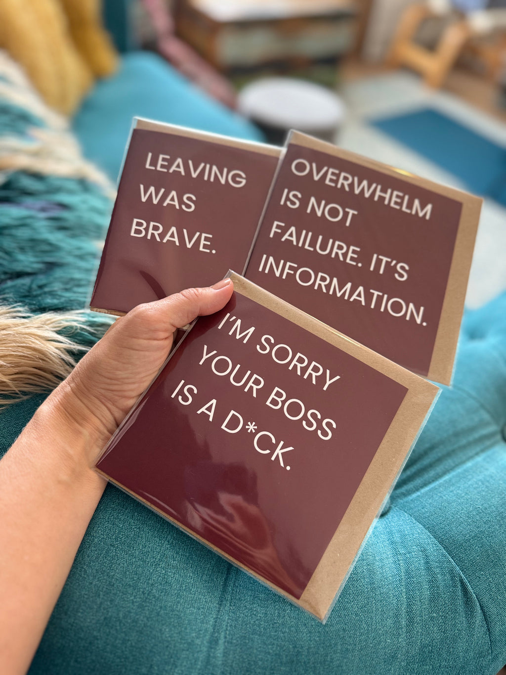 A hand holds three square printed cards with humorous text on a teal couch in a real indoor setting. The cards are dark red with white text and light brown kraft borders.