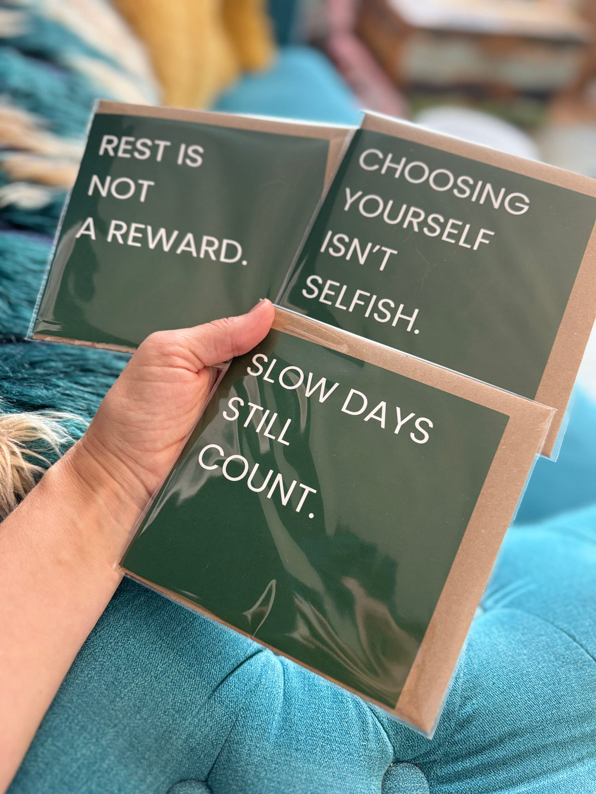 A hand holds three dark green cards with white text that read 'REST IS NOT A REWARD.', 'CHOOSING YOURSELF ISN'T SELFISH. 'SLOW DAYS STILL COUNT>'