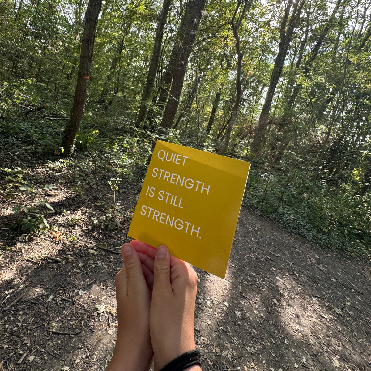 A person holding a gold square card with the text 'QUIET STRENGTH IS STILL STRENGTH.' in white, set against a background of a sun-dappled forest path.