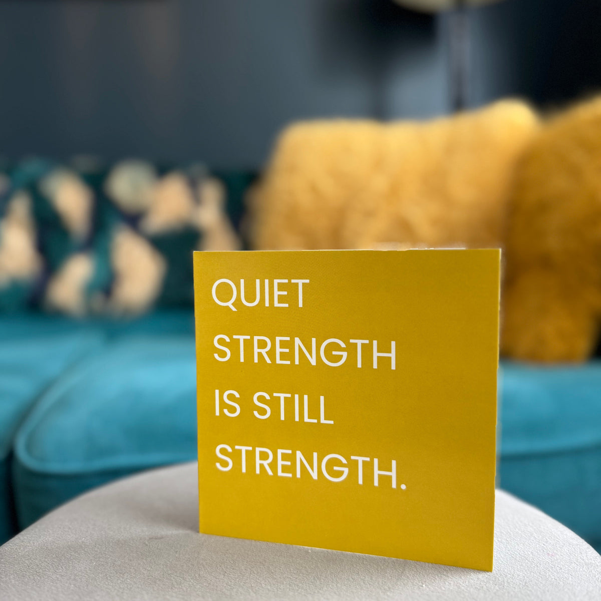 A square yellow sign with white text reading 'QUIET STRENGTH IS STILL STRENGTH.' standing on a light gray surface in a blurred living room setting with a teal sofa and yellow pillows in the background.