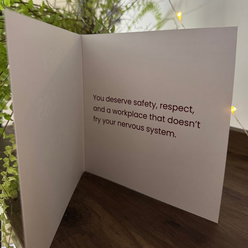 An open white greeting card or letter standing on a wooden surface, with printed text on the inside reading, 'you deserve safety, respect, and a workplace that doesn't try your nervous system.' Green plants and warm string lights are visible in the background.