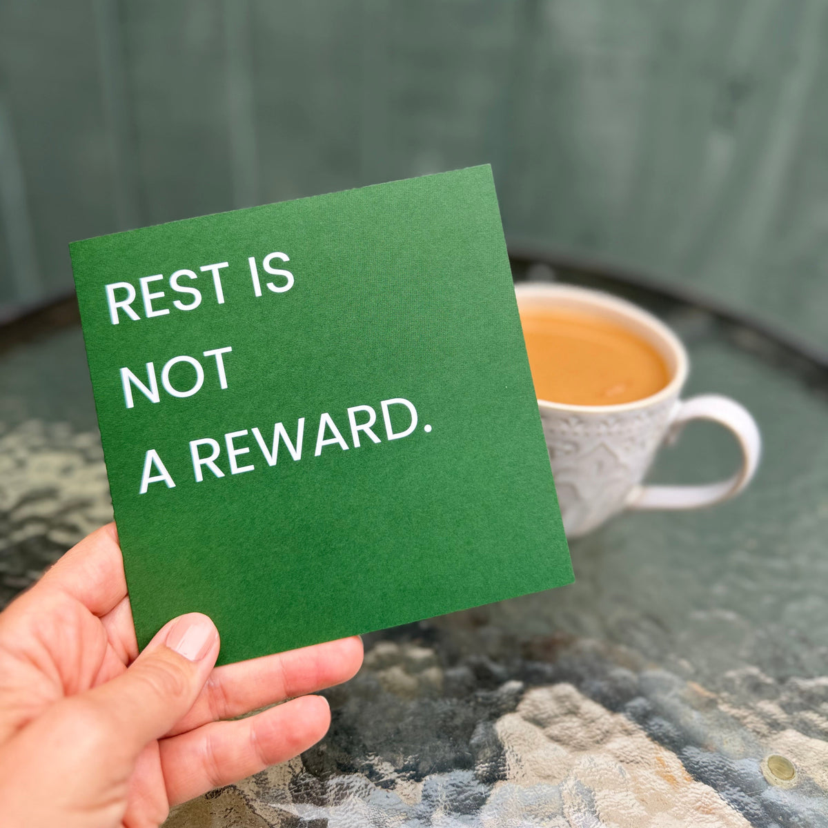 A hand holds a green square card with the white text 'REST IS NOT A REWARD.' in front of a blurred background showing a cup of coffee and a green wooden fence.