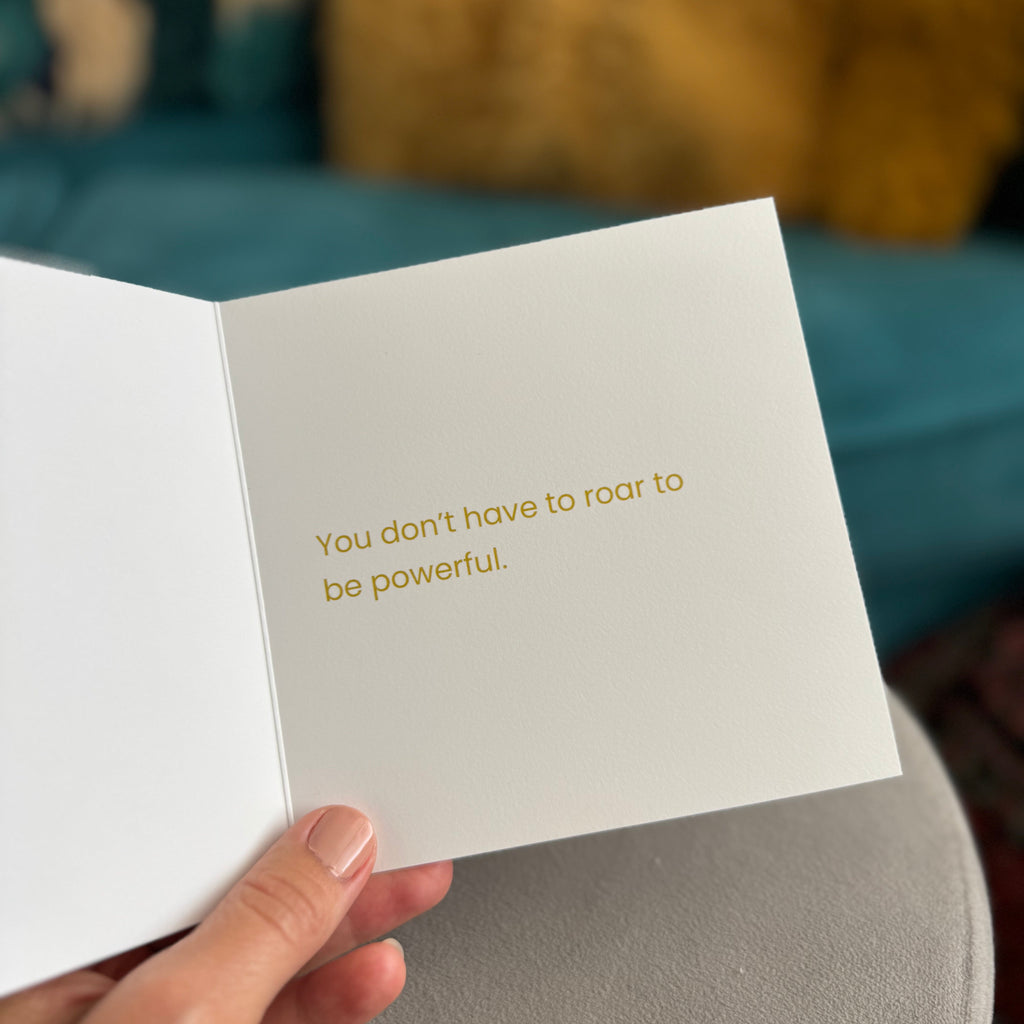 A hand holds an open white greeting card with gold foil text that reads, 'You don't have to roar to be powerful.' The background is a softly blurred living room setting with a teal sofa and yellow pillows.