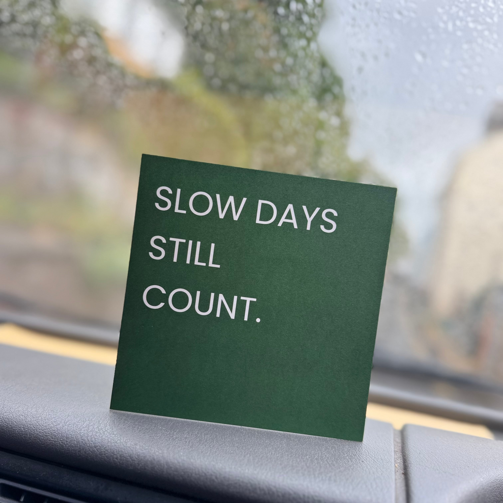 A square dark green sticker with white text reading 'SLOW DAYS STILL COUNT.' placed on a car dashboard in front of a rain-streaked windshield.
