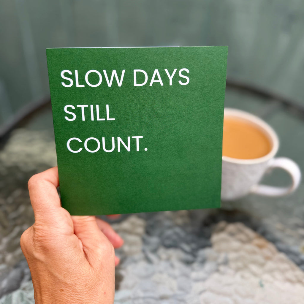 A hand holds a green square card with the white text 'SLOW DAYS STILL COUNT.' in front of a blurred outdoor patio scene with a cup of coffee.