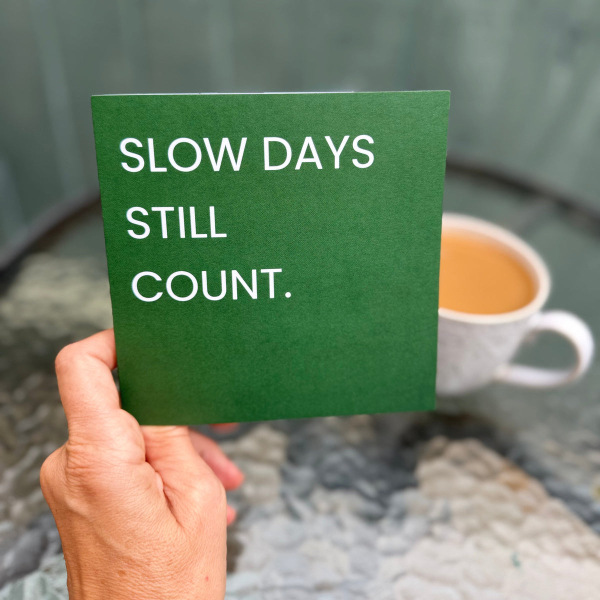 A hand holds a green square card with the white text 'SLOW DAYS STILL COUNT.' in front of a blurred outdoor patio scene with a cup of coffee.