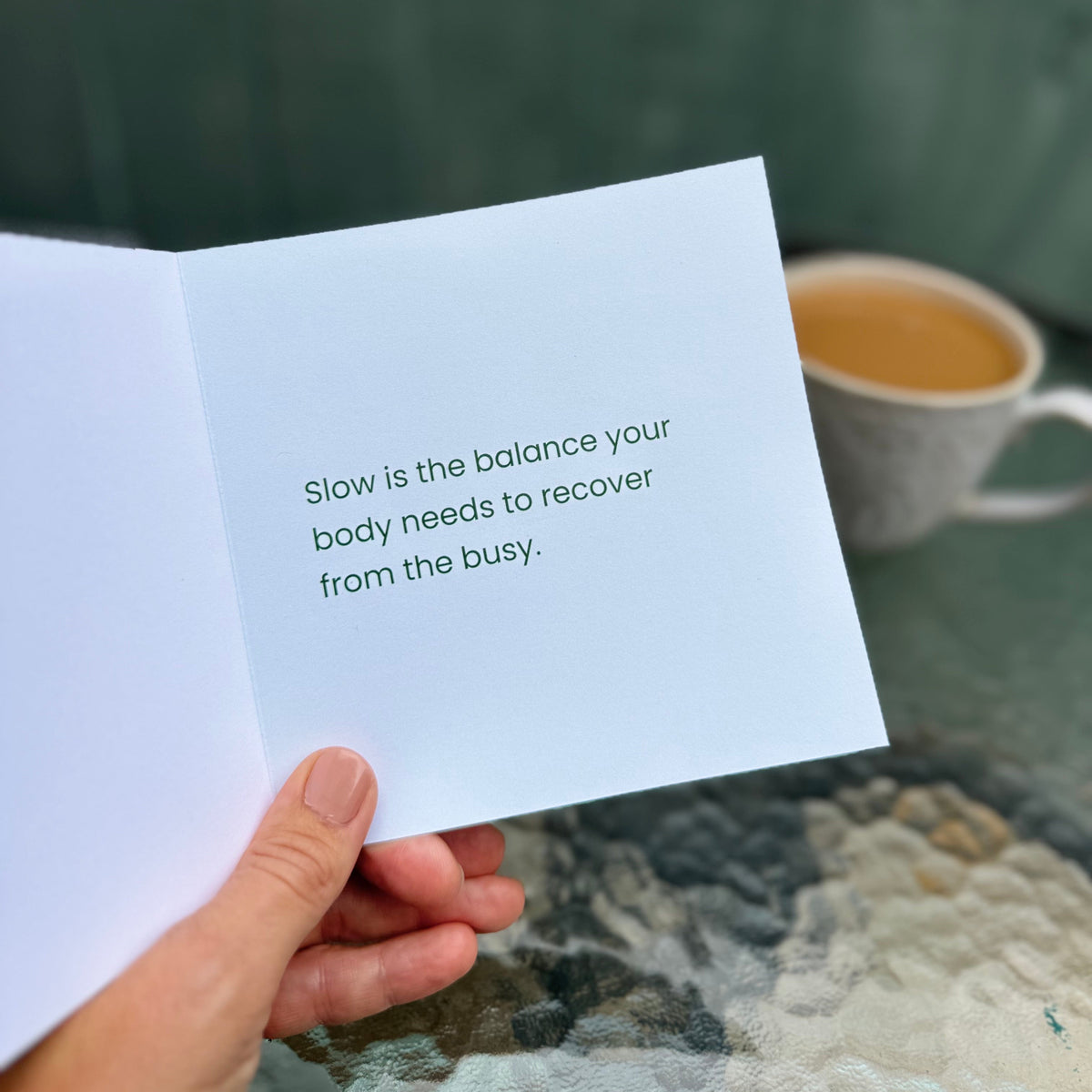 A hand holds an open white greeting card with the printed message, 'Slow is the balance your body needs to recover from the busy.' A cup of coffee sits on a stone table in the blurred background against a dark green wooden fence.