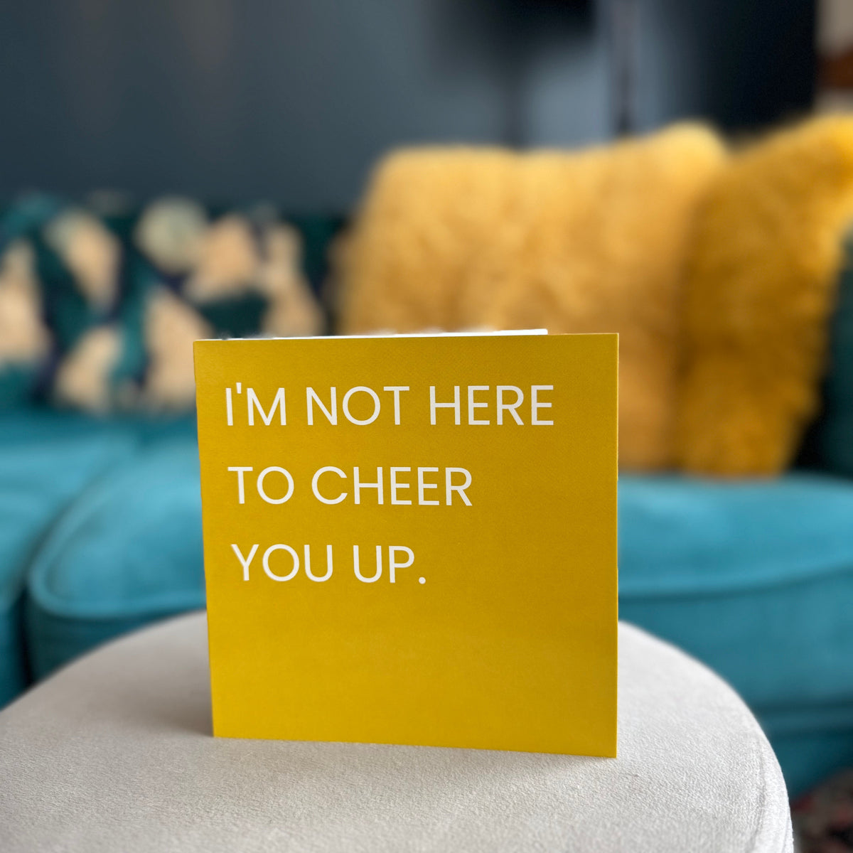 A gold greeting card with white text reading 'I'M NOT HERE TO CHEER YOU UP.' standing on a round white stool in a living room setting with a teal sofa and yellow pillows in the background.
