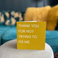 A gold greeting card with white text reading 'THANK YOU FOR NOT TRYING TO FIX ME.' standing on a light-colored round table in a living room setting with a teal sofa and yellow pillows in the background.