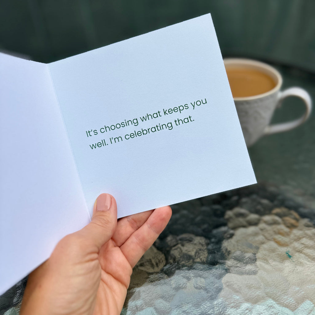 A hand holds an open greeting card with the printed message, 'It's choosing what keeps you well. I'm celebrating that.