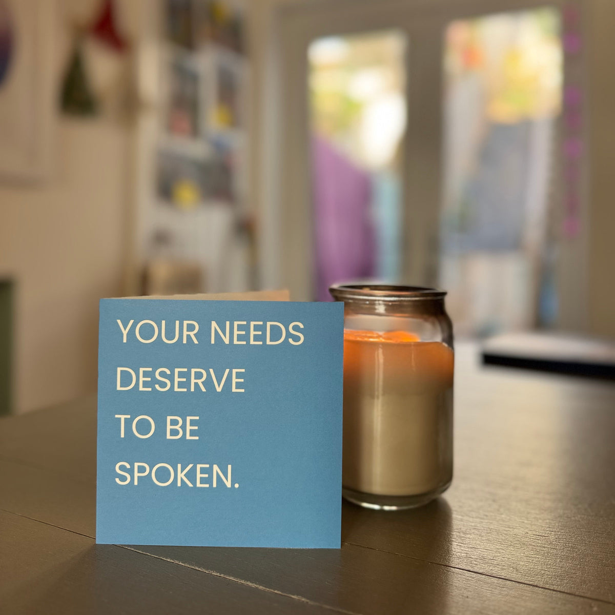 A blue card with the text 'YOUR NEEDS DESERVE TO BE SPOKEN.' sits on a wooden table next to a lit candle in a glass jar, with a softly blurred indoor background.