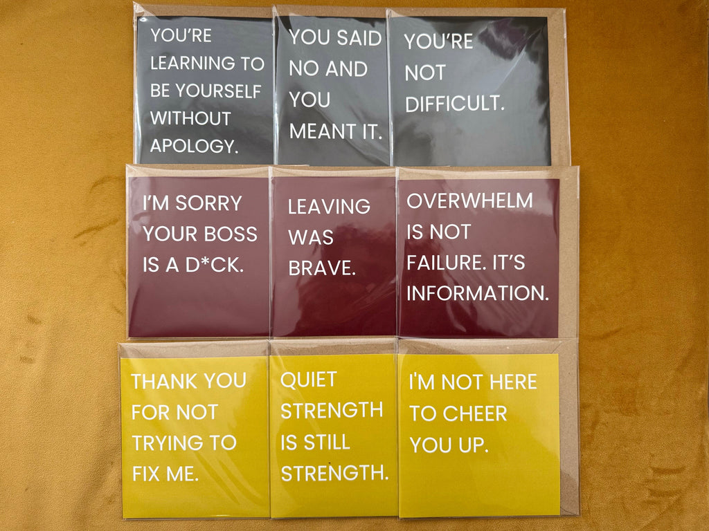 Six square printed cards with motivational or self-care phrases, arranged in a 2x3 grid on a tan fabric background. The cards come in black, red, and yellow, each with white text.