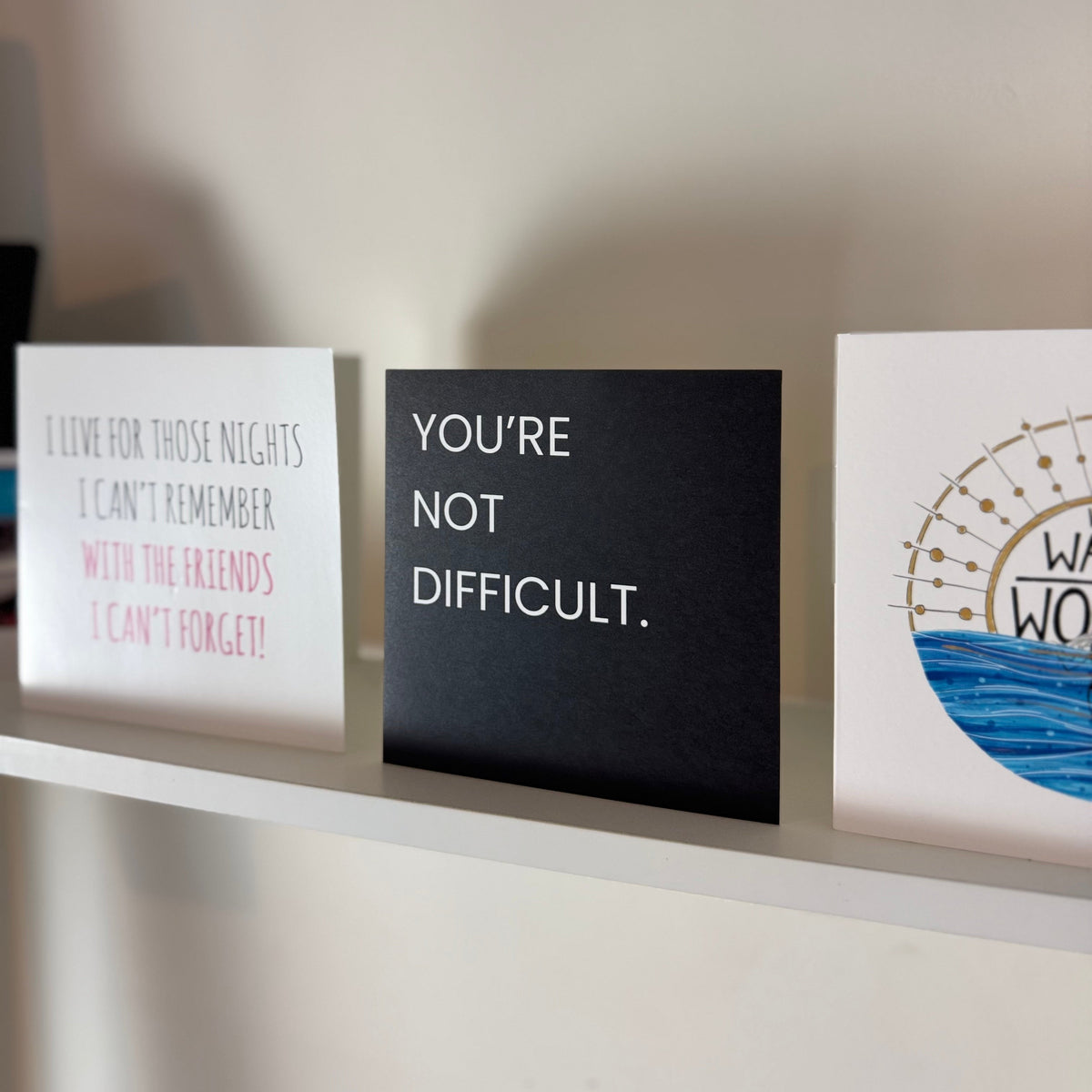 Decorative wall art cards displayed on a white shelf against a beige wall. The center card is black with white text reading 'YOU'RE NOT DIFFICULT.