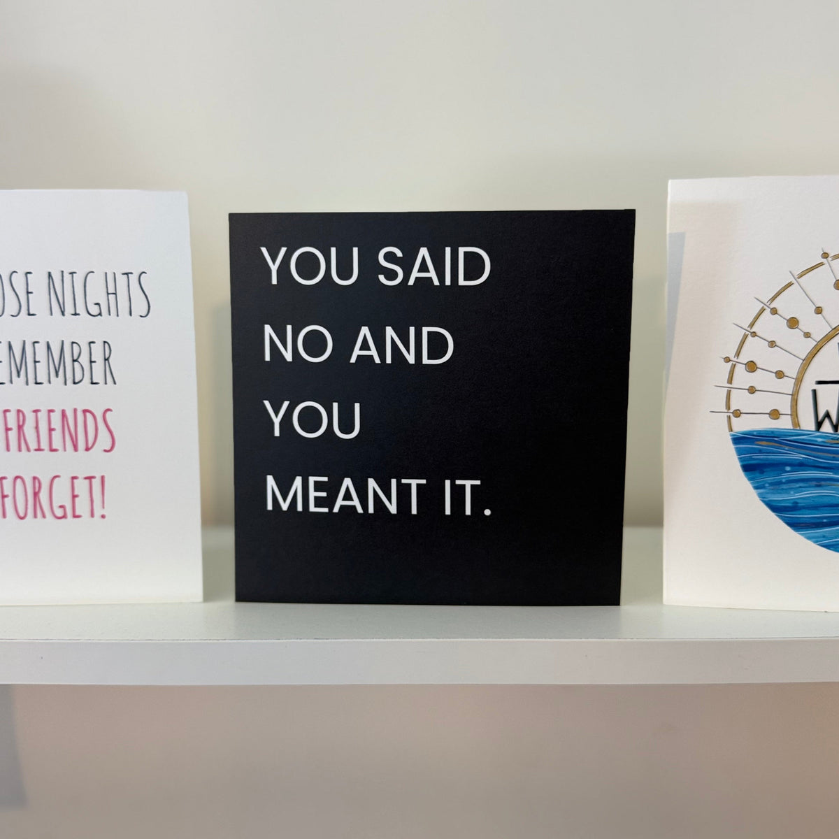 Three typographic art cards displayed on a white shelf against a light gray wall. The center card is black with white text reading 'YOU SAID NO AND YOU MEANT IT.